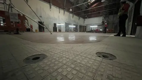 Close up of Jacobs Wells Baths' current flooring inside with square tiles. Scaffolding has been erected inside and there are workmen in the background.