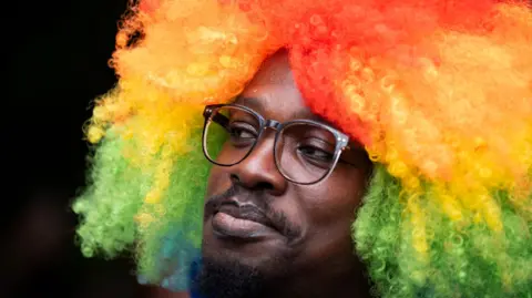 PA Media Close-up portrait of a Pride attendee wearing glasses and a large curly rainbow-coloured wig with red, orange, yellow, green and blue sections.