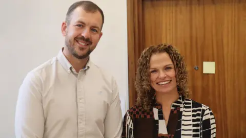 Slack, wearing a white shirt , stands next to Clarke. 