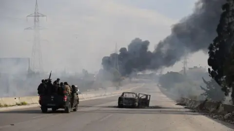 Abdulvacit Haci Isteyfi/Anadolu via Getty Images A group of about eight men in military clothing ride on a jeep, with the barrel of a gun pointing into the air above them. The jeep is driving towards clouds of thick dark smoke, along a road that is empty apart from one damaged car stuck in the middle of it.