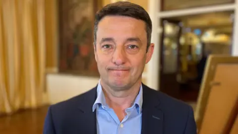 Boley Smillie - A man with black hair looking towards the camera. He's wearing a blue suit jacket and a blue shirt. 