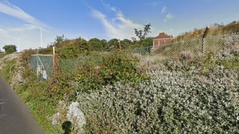 A view of Eastney East Battery which is obstructed by bushes and shrubs. The area is fenced off.