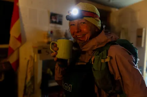 Adam Wild Gossage, wearing a purple coat, yellow hat and a torch on her head, stops for a hot drink at one of the checkpoints along the route. 