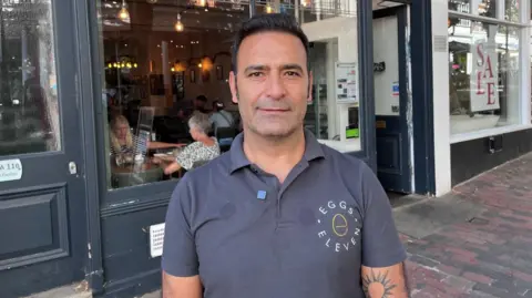 Gurky Koca, owner of Eggs Eleven cafe in the Pantiles, Tunbridge Wells. Gurky is stood outside the cafe. He is wearing a t-shirt with the 'Eggs Eleven' logo. He has tanned skin and black hair.