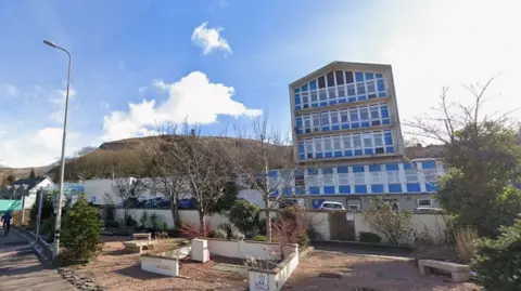 Picture of the outside of Belford Hospital, a multi-storey building painted blue and white. There is a hill in the background, and it is a sunny day with some clouds in the sky.