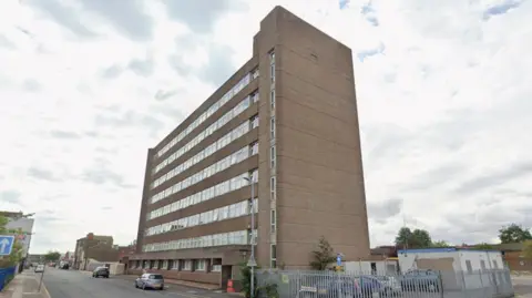 A large, multi-storey office building. Cars are parked on the street outside the building. The sky is cloudy.