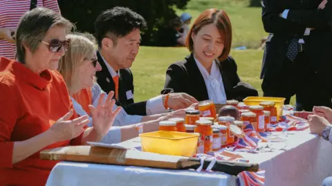 Mary-Lee Winkler People sit around an outdoor table lined with more than a dozen jars of orange-coloured marmalade. The four people on the left are smiling and concentrating on the jars before them. There are people stood behind the, The table also has union jack flag bunting on it and yellow plastic tubs.