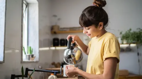 A woman in a yellow tshirt stands at a kitchen sink, pouring water from a silver stovetop kettle into a mug.