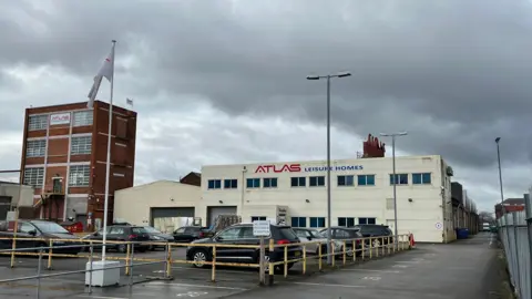 The exterior of the Atlas Leisure Homes headquarters on Wiltshire Road in Hull. The main building is covered in cream cladding and has a large blue and white logo spanning the width of the building. A building on the left is a red brick tower block with six large windows. A car park is in front of the two buildings, along with a flagpole and lights.