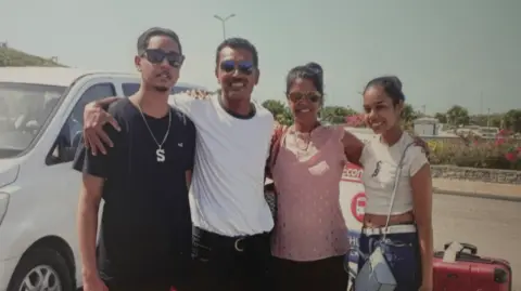 Shawn Seesahai family with his parents and sister; They are all smiling at the camera and have their arms around each other. They are standing outside and behind them is a vehicle to their left and a suitcase to their right