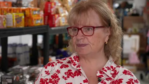 Penny wears glasses and a white shirt with red flowers on it.