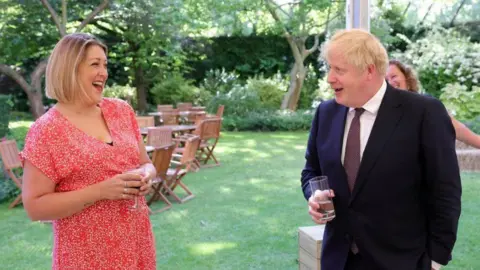 No 10 Downing Street Holly King-Mand is talking to Prime Minister Boris Johnson in the garden at No 10 Downing Street in 2021. She has blond shoulder-length hair and is wearing a red dress with a white pattern and is looking at the prime minister and laughing.  Boris Johnson, who has bright blond hair is wearing a dark suit, white shirt and maroon tie. He is looking at Ms King-Mand and is smiling. Both and holding glasses with liquid in them.