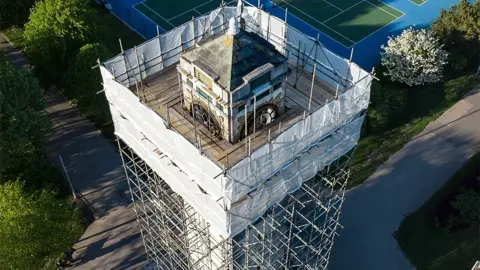 Drone view of the Cocker Memorial Clock Tower covered in scaffolding, with white sheeting to the very top. In the background are gardens, paths and tennis courts