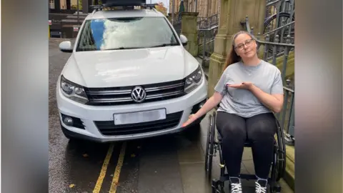 Jennie, a woman in a wheelchair, gesturing towards a white Volkswagen family car which has been parked on a pavement, blocking her from being able to get past without travelling in the road itself.