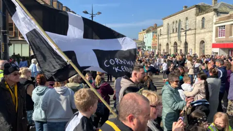 The black Cornish flag with a white cross superimposed on it is held aloft at a St Piran's Day gathering in Lemon Quay in Truro. A large crowd is gathered and it is a sunny day.