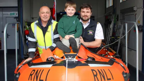 Southend RNLI A granddad and a dad sat in an orange RNLI lifeboat with the grandson sat on top of it