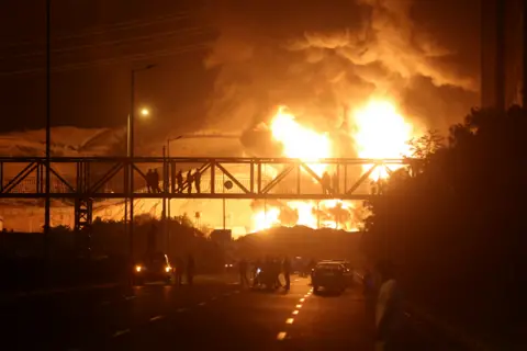 Reuters People watch from a bridge as flames from an Israeli attack rise from Sharan Oil depot