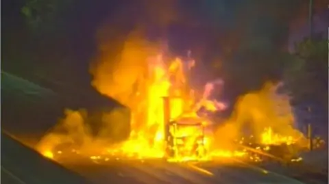 National Highways A lorry on fire on the M25, showing a burnout vehicle, the rest of the image is blurred. 