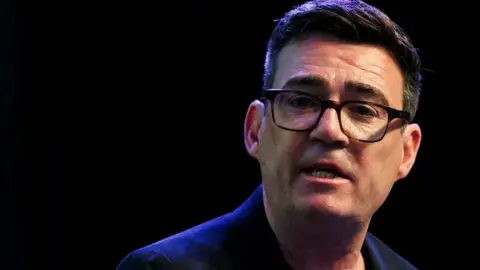 Andy Burnham has dark hair, dark spectacles and wears a dark blue jacket.