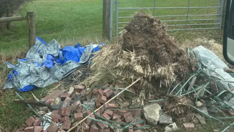 Steven Conisbee Bricks, sheeting and garden waste are dumped on farmland. There is a gate behind the waste looking out onto a field.