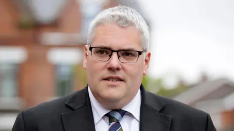 PA Media Gavin Robinson pictured outside wearing a grey suit, green tie and spectacles. 