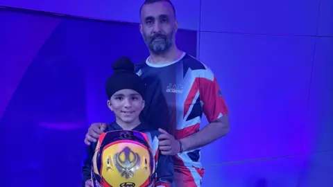 A young boy holding a karting helmet with a sign of the Sikh religion. He is pictured with his dad placing both hands on his shoulders. 