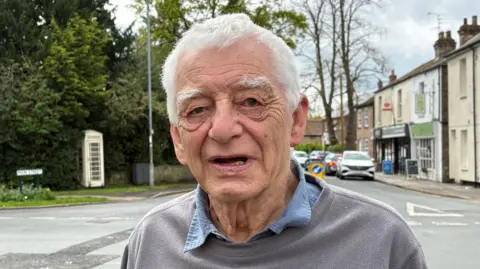 Kevin Shoesmith/BBC A man in his 80s, with short grey hair, poses for the camera. He is wearing a blue shirt under a grey sweatshirt. Behind him is a parade of village shops, including a convenience store. 