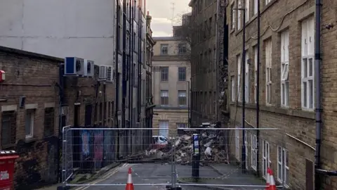 Charles Heslett/BBC Rubble in a pile blocking a road with a metal barrier fence and orange and white cones cordoning off the scene. There are buildings on both sides of the road. 