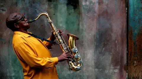 A musician leaning back as he plays on a saxophone. He is wearing a brown leather cap and a mustard coloured shirt. Behind him is a concrete wall.