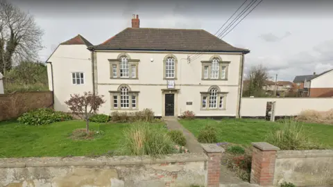 A street view image of a grand-looking beige building with grass and trees in front of it.