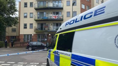A police van outside a block of flats, with a police cordon around the building and two officers outside