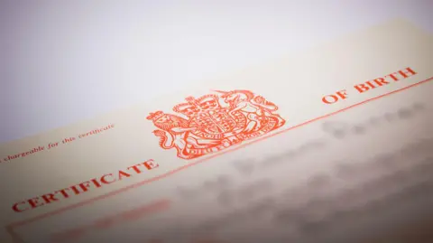 Getty Images A UK birth certificate is on a white background. The paper is off-white with red font. It says Certificate of Birth and has a red logo in the centre. 