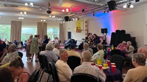 Lily-May Symonds/BBC A room full of people sitting around tables with red plastic cups and bottles of lemonade. A crowd is also on the dancefloor.