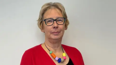 A woman with short blonde hair, glasses, a multi-coloured necklace, a black top and a red cardigan, stands in front of a white wall and smiles.