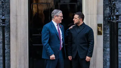 Getty Images A bespectacled man in a blue suit, white shirt and purple tie pats the shoulder of a bearded man in a black jacket and shirt in a doorway.