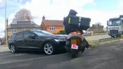 Moped with L plates at road junction with dark blue unmarked police car boxing in the moped to stop it.