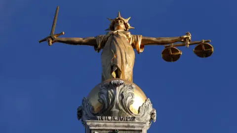 A close up of the statue of Justice that sits at the top of the Old Bailey court building. The statue is of a robed woman with a sword in one hand and a scales in the other. The golden statue stands against a clear blue sky. 