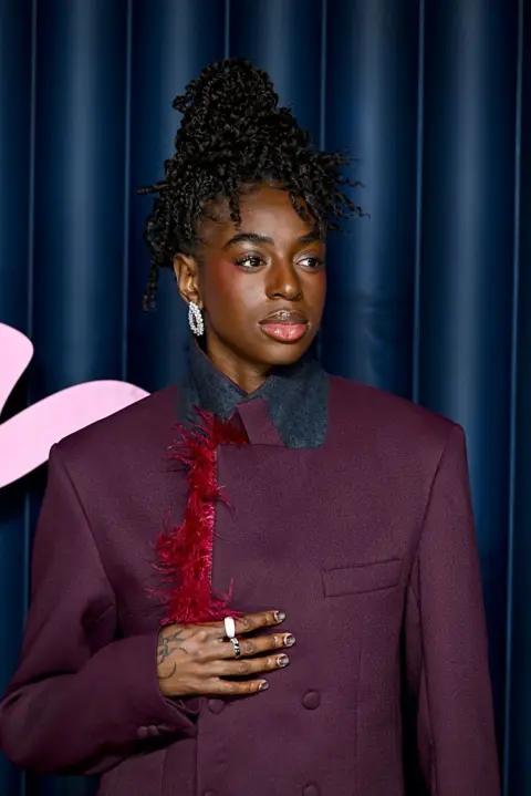 Getty Cat Burns looks off to the side, wearing a purple suit fringed with red feathers