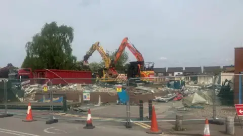 A building site fenced off and surrounded with traffic cones. An orange and yellow bulldozers are positioned on top of a flattened building. Houses can be seen in the background. Bulldozers demolishing a condemned hospital building