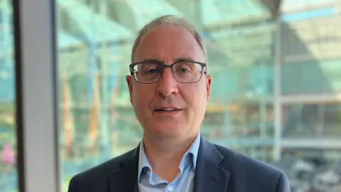 Alex Dunlop/BBC A man with glasses wearing a suit and shirt looks towards the camera