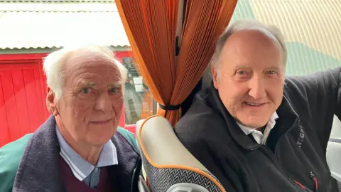 Rod Dolby, a man with white hair, is wearing a green coat with a black collar. He is sitting behind John Bradshaw on a coach. John has grey hair and is wearing a black zipped-jumper. They are both smiling. 