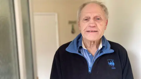 An elderly man with grey hair is wearing a black and blue fleece and polo shirt. He is smiling into the camera and stood at a front door.