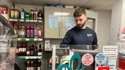 James Casinelli stands behind the cash register in the store. He looks down as he stands in front of the bottles on the shelves behind him. 