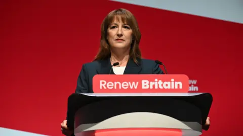 Rachel Reeves stojí na pódiu před červeným pozadím. Na pódiu se říká "Obnovit Británii".