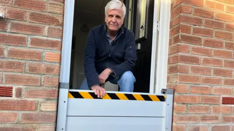 BBC A man crouches behind a metal flood barrier at his front door.