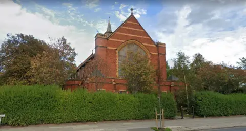 Image shows the red brick St Andrews church in Bootle. It stands behind a large hedge on a main road. 