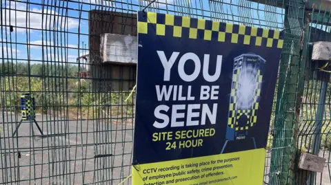 Mid Suffolk District Council A yellow and blue sign that reads "You will be seen Site Secured 24 Hour". It is hanging on a green fence that surrounds a piece of land. 