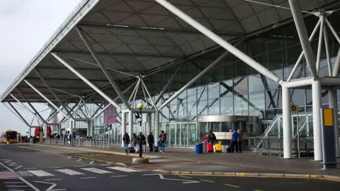 A general view of the express set down at Stansted Airport. There are two lanes in front of the large terminal building, which has a glass facade and large grey columns propping up its flat roof.