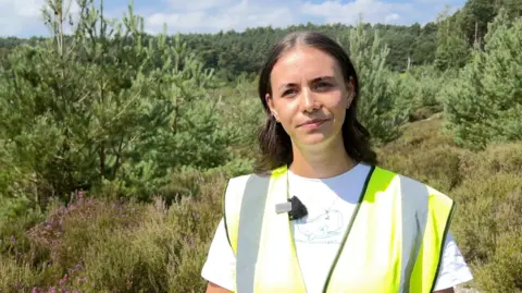Izzy Bridges, ecologist for the Ministry of Defence, wearing a hi-viz waistcoat over a white t-shirt. In the background there are green trees and small purple flowers.