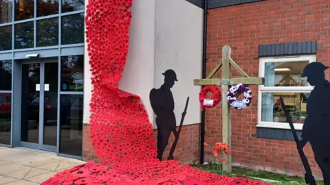 Avery Healthcare A stream of poppies fixed to the side of a building's entrance with two soldier silhouettes and a wooden cross beside it